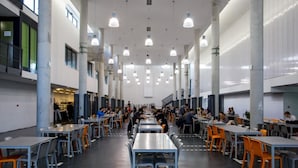 Eine Cafeteria mit mehreren Tischen und Stühlen, großen Säulen und hängenden Lampen.