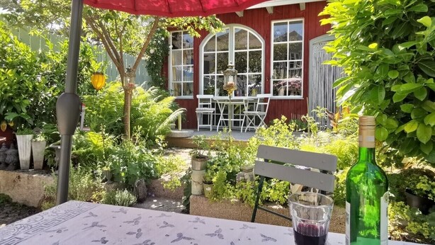 Eine Außenpatio mit Tisch, einer Weinflasche und einem Glas. Ein roter Sonnenschirm, eine weiße Bank und ein rot gefasstes Gebäude mit großen Fenstern sind im Hintergrund zu sehen.