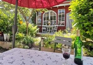 Een terras buiten met een tafel, een fles wijn en een glas. Er is een rode parasol, een witte bank en een rood gebouw met grote ramen op de achtergrond.