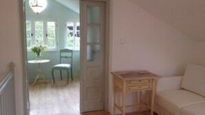 Ein Zimmer mit Holzboden, einem weißen Sofa, einem kleinen Holztisch mit Schubladen, einem Sessel und einer Tür zu einem weiteren Raum mit Fenster und einer Chandelier.