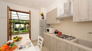 A modern kitchen with wooden cabinets, a gas stove, a microwave, and a refrigerator. There is a dining table with chairs and a small garden visible through an open door.