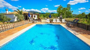 Ein rechteckiger Pool mit Fliesenrand, umgeben von Liegestühlen und Sonnenschirmen, vor einer Kulisse aus Bäumen und Hügeln.