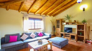 Ein gemütliches Wohnzimmer mit Holzbalkendecke, einer Couch mit Kissen, einem Fernseher und einer Kommode mit Dekoration.