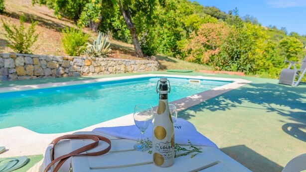 A bottle of Lolei Brut champagne, a glass, and a brown belt on a table by a pool with a stone wall and greenery in the background. - Casa Rural Molino de la Máquina Cartajima by Ruralidays (Málaga)