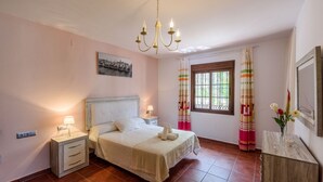 Une chambre à coucher avec un lit, des tables de chevet, un lustre, une fenêtre avec des rideaux et un tableau au mur.