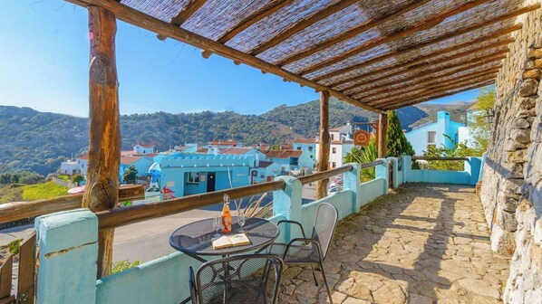A terrace with a table and chairs, overlooking a village with blue buildings and a mountainous backdrop.