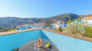 A rooftop infinity pool with a bottle and glasses, overlooking a village with terraced houses and hills.