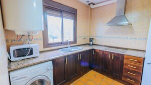 A compact kitchen with a washing machine, microwave, sink, and wooden cabinets. - Casa Genal Júzcar by Ruralidays (Málaga)