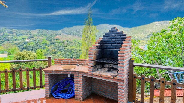 A brick-built outdoor grill with a chimney, surrounded by a wooden fence and a view of a lush green landscape. - Casa Genal Júzcar by Ruralidays (Málaga)