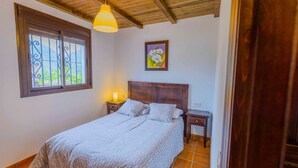 A bedroom with a bed, a nightstand, a lamp, a window with a view, and a painting on the wall. - Casa Genal Júzcar by Ruralidays (Málaga)