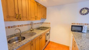 A kitchen with wooden cabinets, a granite countertop, a sink, a microwave, and a wall clock. - Casa Diego Piña Júzcar by Ruralidays (Málaga)