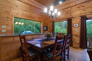 Interno di una baita in legno con tavolo e sedie, un lampadario e ampie finestre che danno sulla foresta.