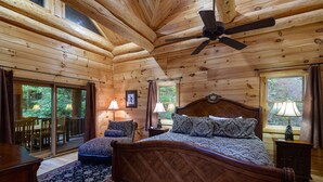 A wooden cabin bedroom with a bed, two lamps, a ceiling fan, and a view of the outdoors.