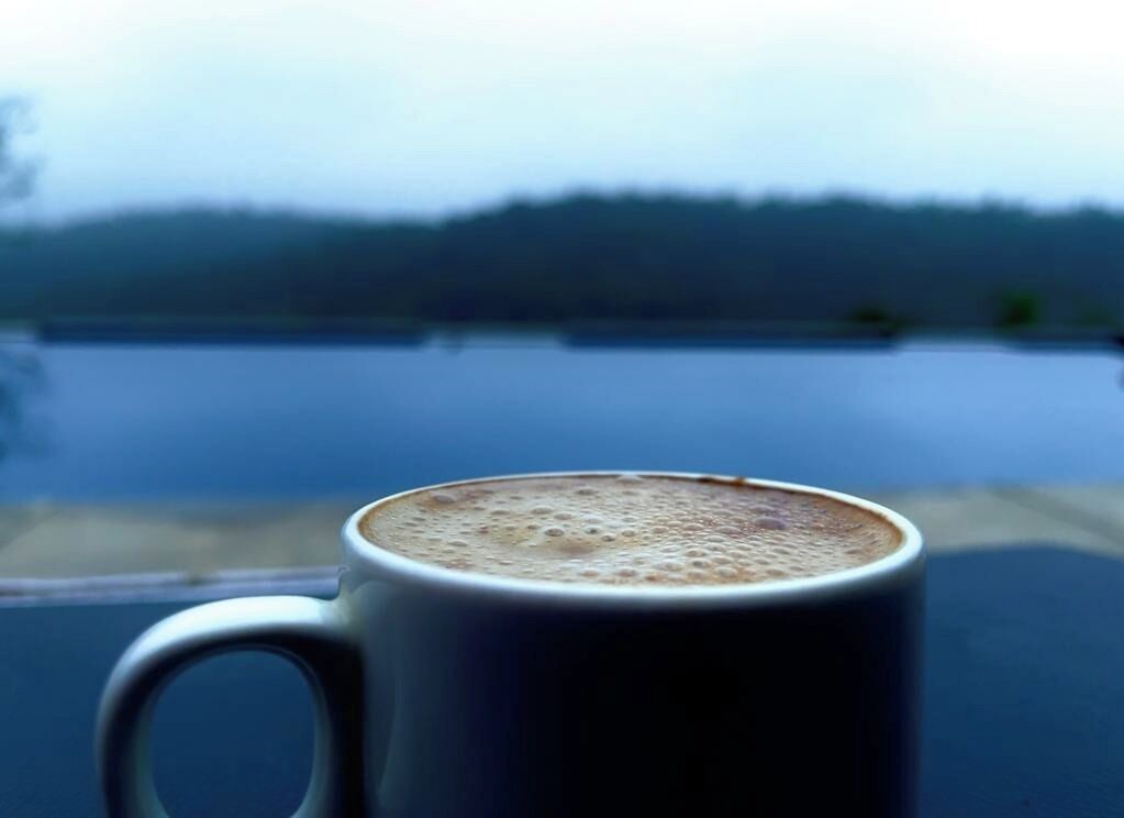 A close-up of a coffee mug with a frothy top, placed on a table with a view of a calm lake and distant trees.
