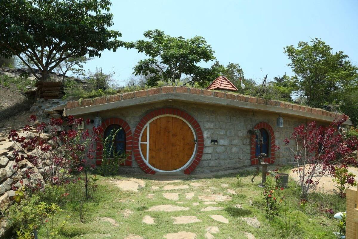 A stone house with a wooden door and two arched windows, surrounded by a garden with red-leaved plants.