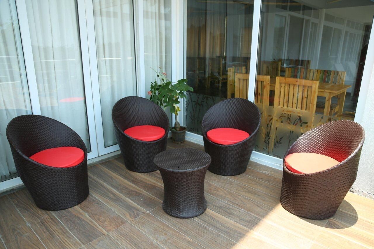 A balcony with wicker chairs and a small table, a potted plant, and a view of a wooden deck and chairs through a glass door.