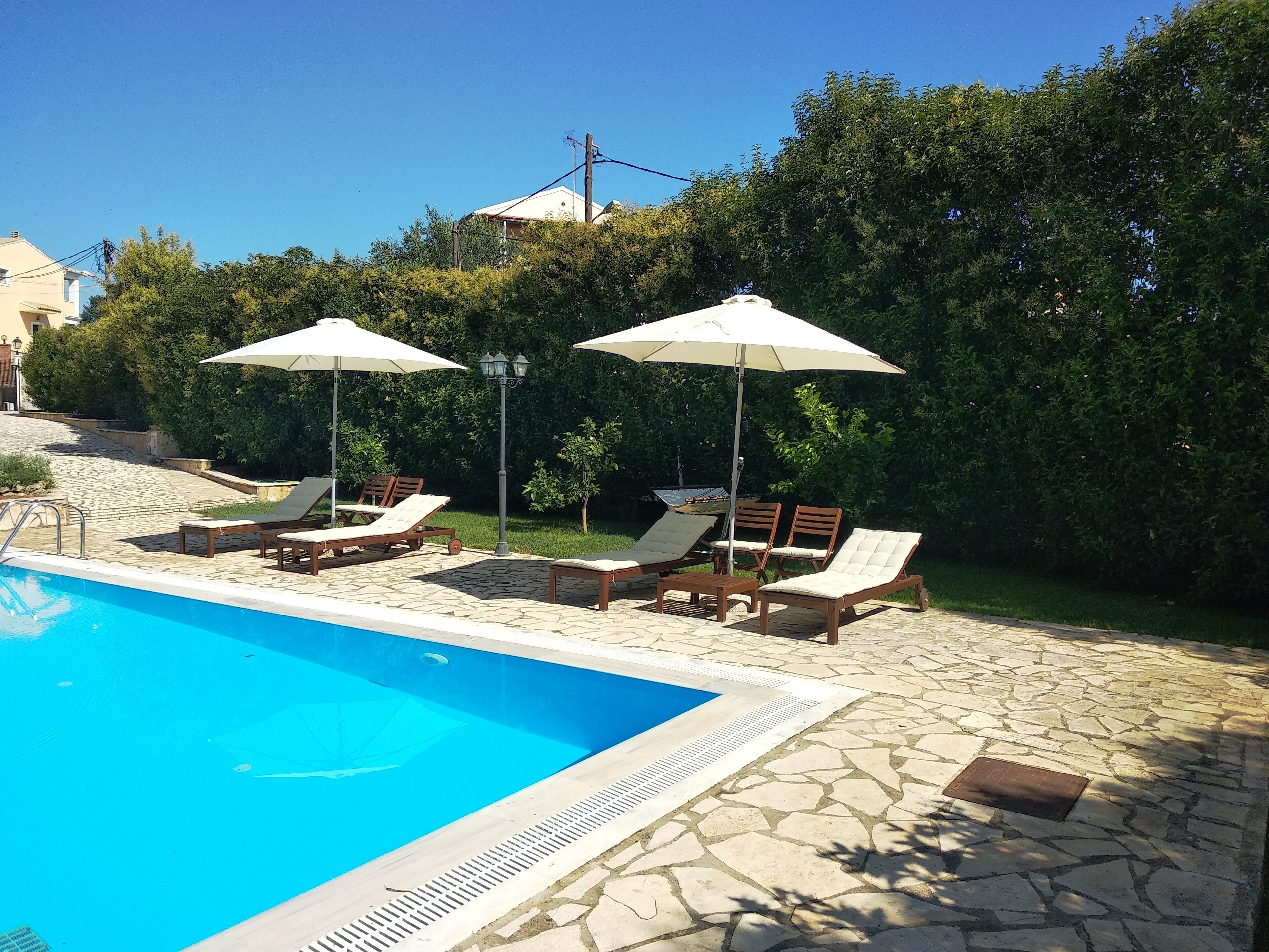 Una piscina con sillones de descanso y sombrillas, rodeada de baldosas de piedra y follaje.