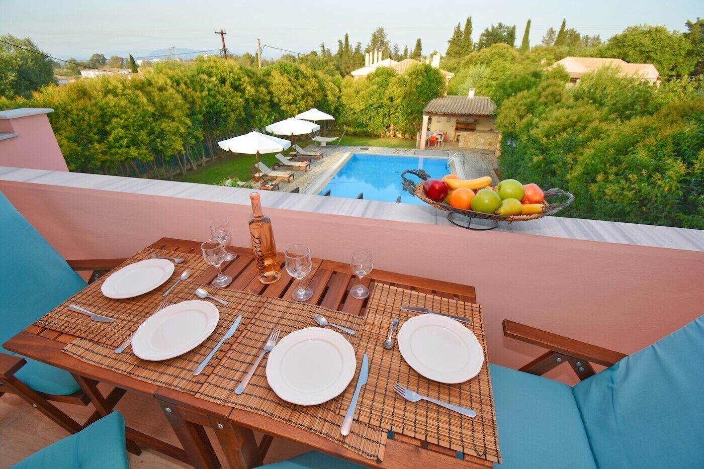 A dining table set with plates, cutlery, and a bottle, overlooking a swimming pool and lush greenery.