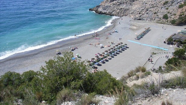 Una playa con hileras de sombrillas y tumbonas, acantilados rocosos y el mar.