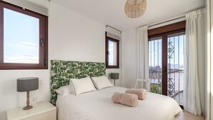 Un dormitorio con una cama grande, dos lámparas de noche, una mesita de noche y vistas de la ciudad desde un balcón con barandilla.