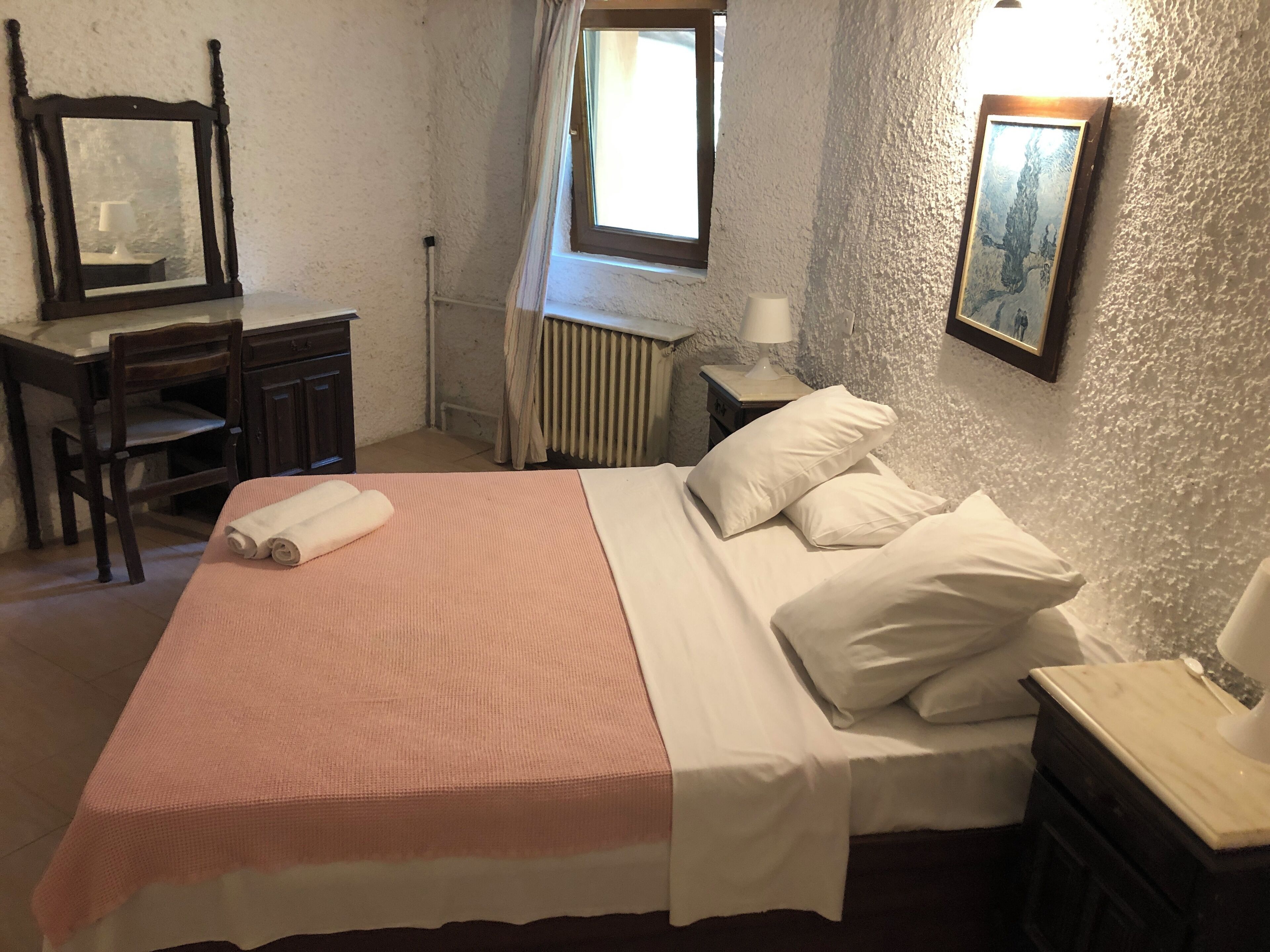 A neatly made bed with white linens and a pink blanket, a wooden chair, a desk with a mirror, a radiator, and a wall-mounted picture.