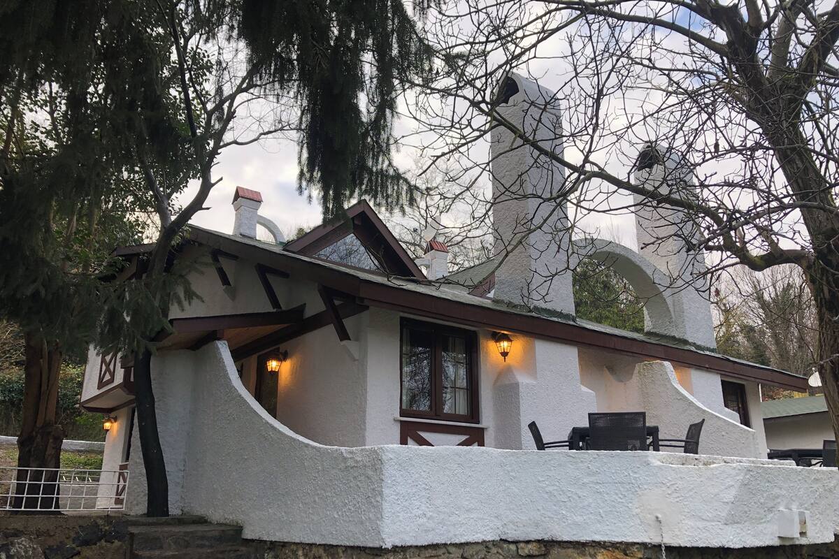 A rustic cabin with a stone foundation, a sloped roof, and a chimney. There is a porch with outdoor seating and a lantern.