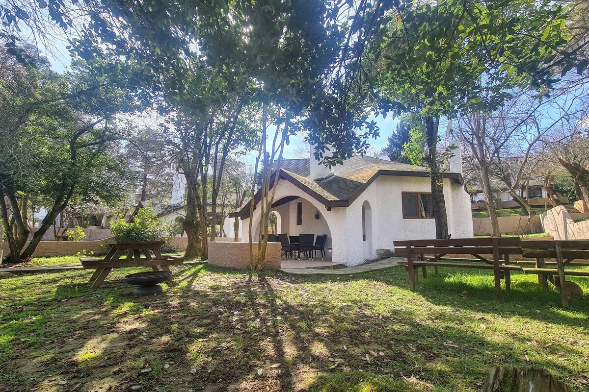 A rustic building with a white exterior, surrounded by trees and a grassy area. There are picnic tables and a small patio with chairs.