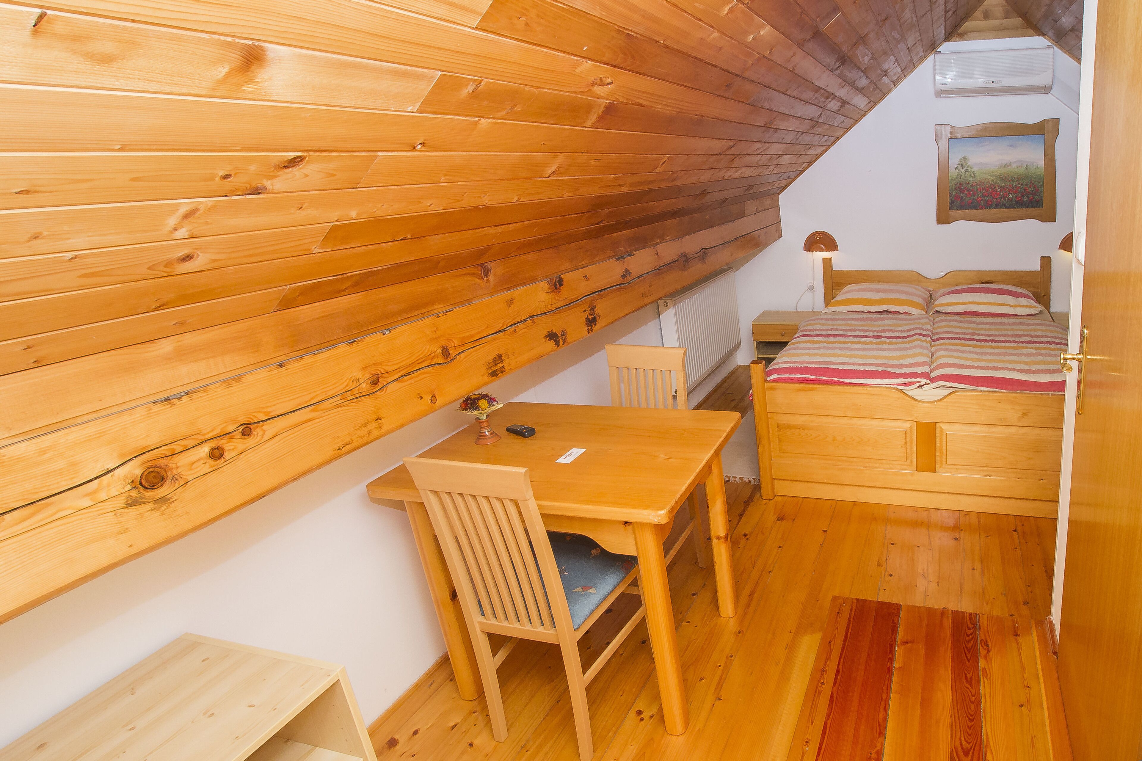 A small, cozy room with wooden walls and a sloped ceiling. It includes a bed, a desk, and a chair.