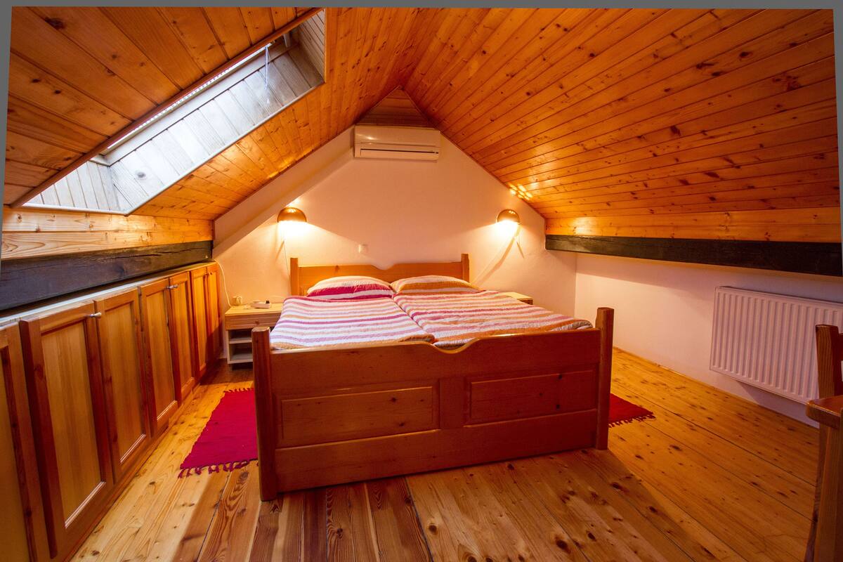 A cozy attic bedroom with a wooden ceiling, a bed, a nightstand, and a red rug.