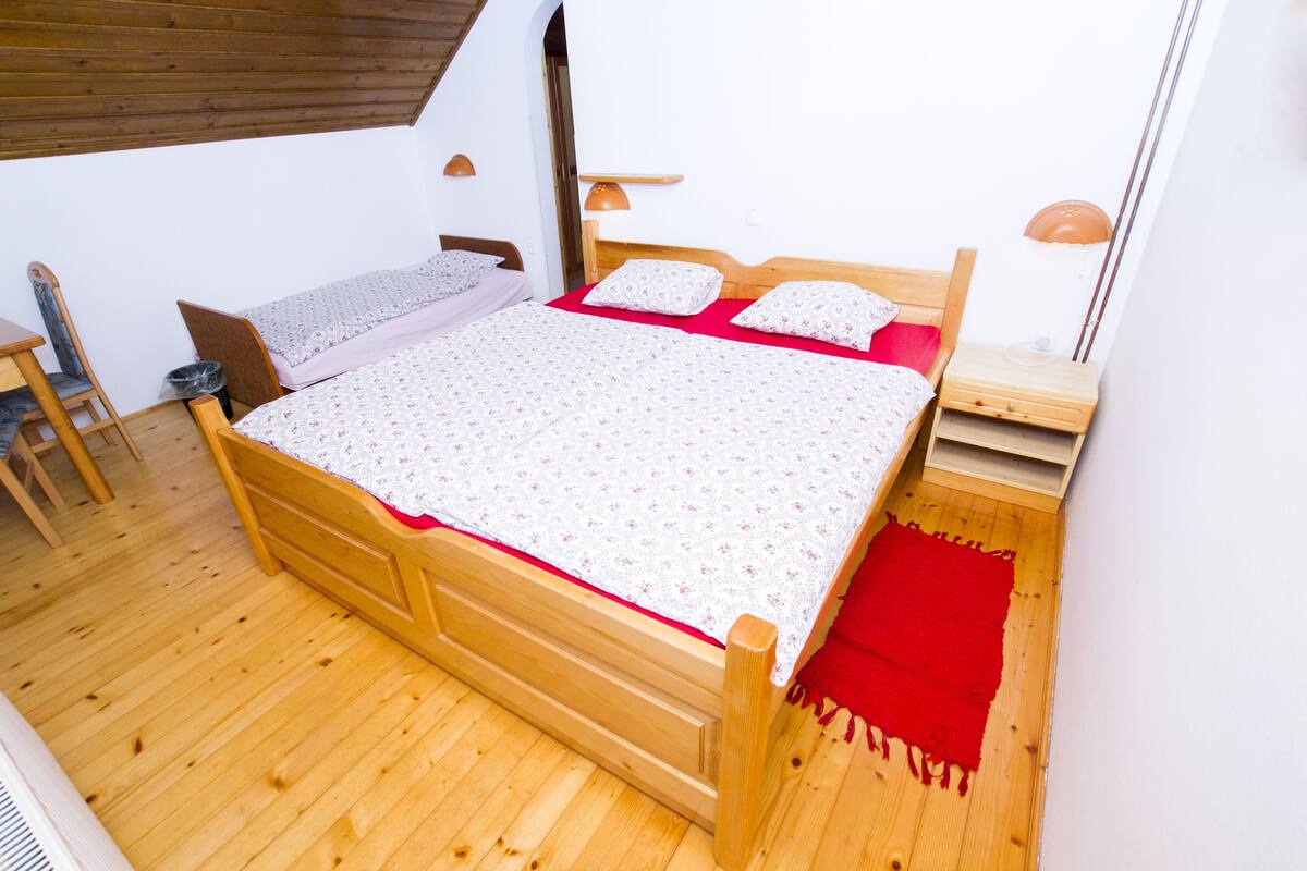 A wooden bed with a red blanket, a wooden nightstand, and a chair in a room with wooden flooring and white walls.