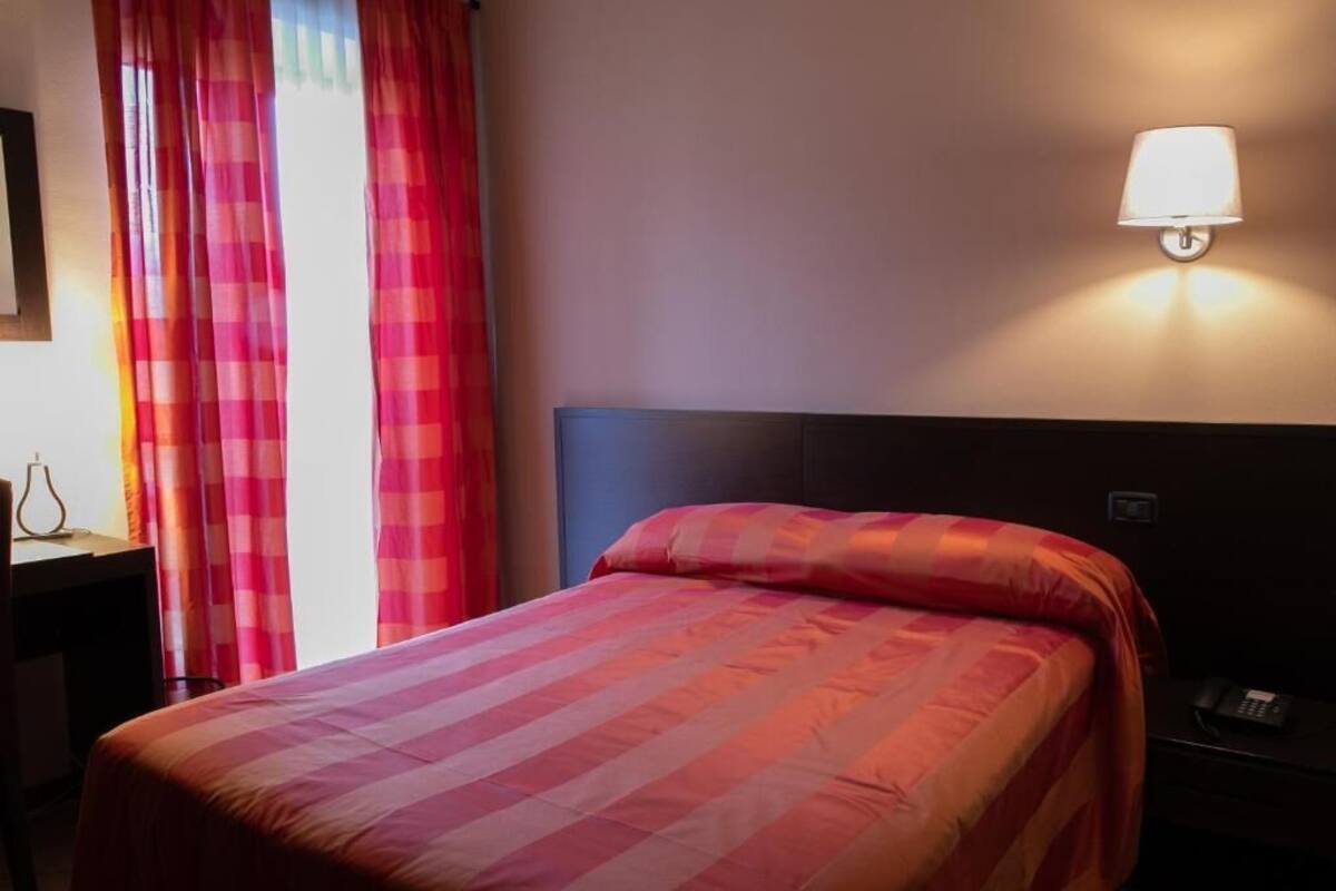 A hotel room with a bed, a nightstand, a lamp, and a window with curtains.