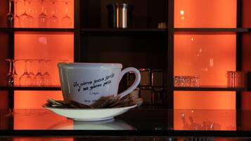 Eine weiße Keramiktasse mit Schriftzug auf einem Untertasse, platziert auf einer reflektierenden Oberfläche mit Regalen im Hintergrund.