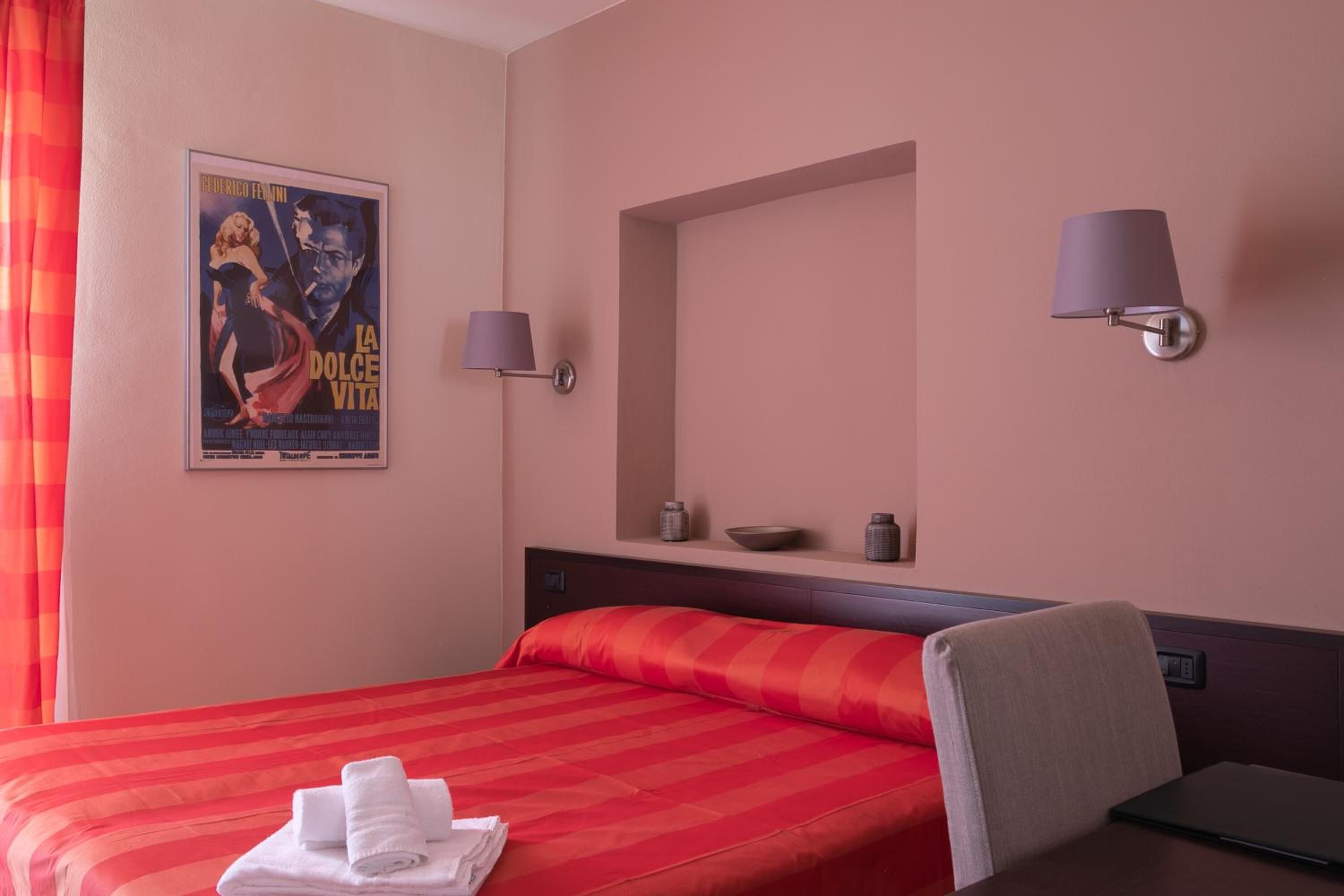 A hotel room with a large bed, a chair, a desk, a poster on the wall, and two wall-mounted lamps.