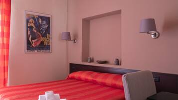 A hotel room with a large bed, a chair, a desk, a poster on the wall, and two wall-mounted lamps.