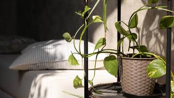 A wicker plant stand with a potted plant on a wicker table, positioned in a room with a bed and a pillow in the background.