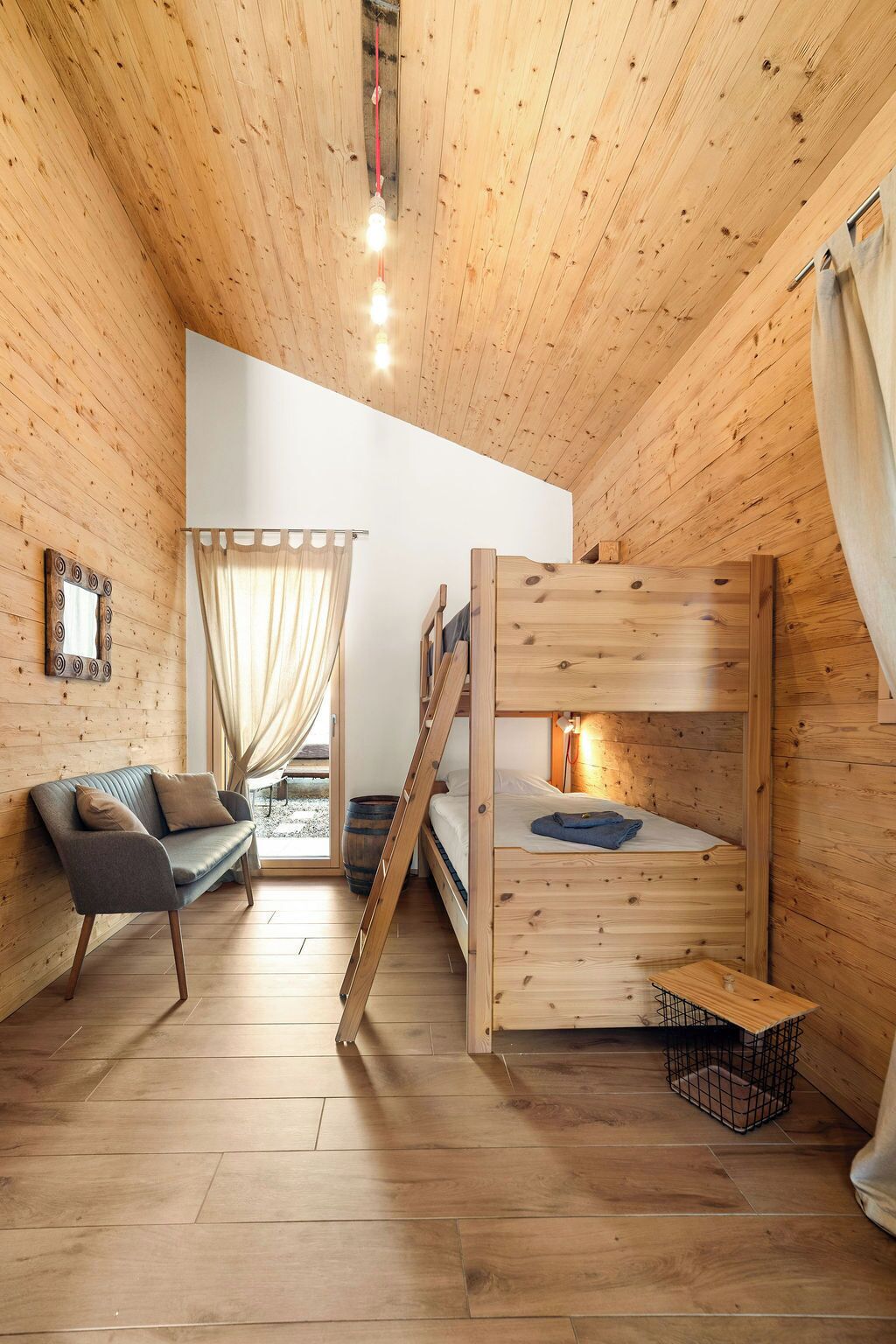 A wooden cabin interior with a bunk bed, a sofa, a mirror, and a window with curtains.