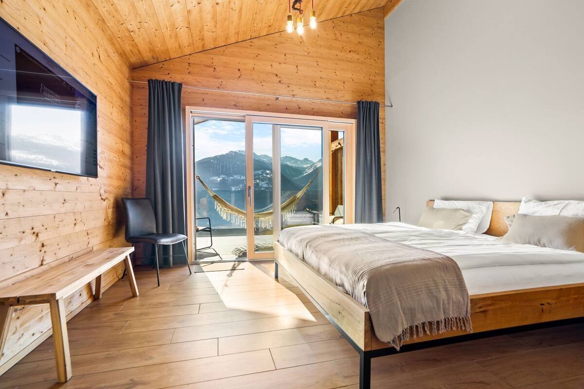 Une chambre dans une cabane en bois, avec un lit, un banc, une chaise et un hamac près d’une porte-fenêtre coulissante.