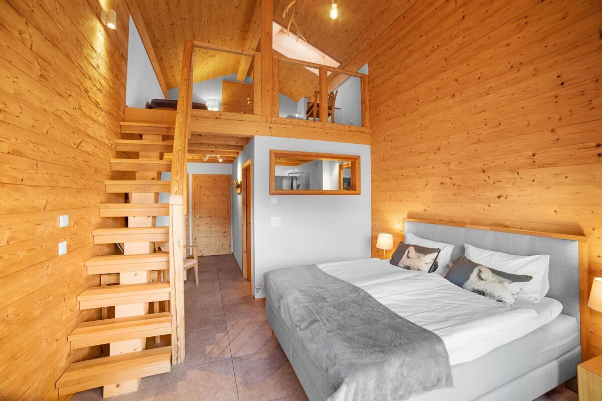 Une chambre aménagée dans une cabane en bois, avec un lit, une table de chevet, un miroir et un escalier menant à un étage supérieur.