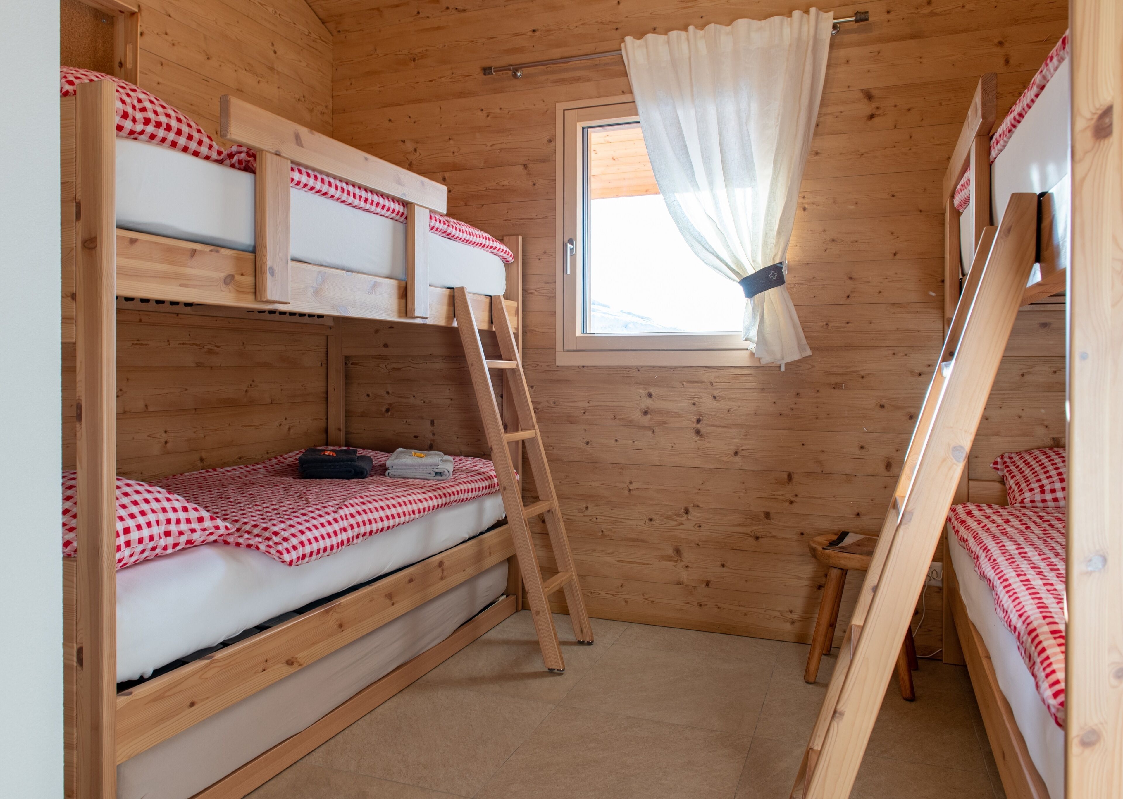 A wooden cabin room with bunk beds, a ladder, and a window with curtains.