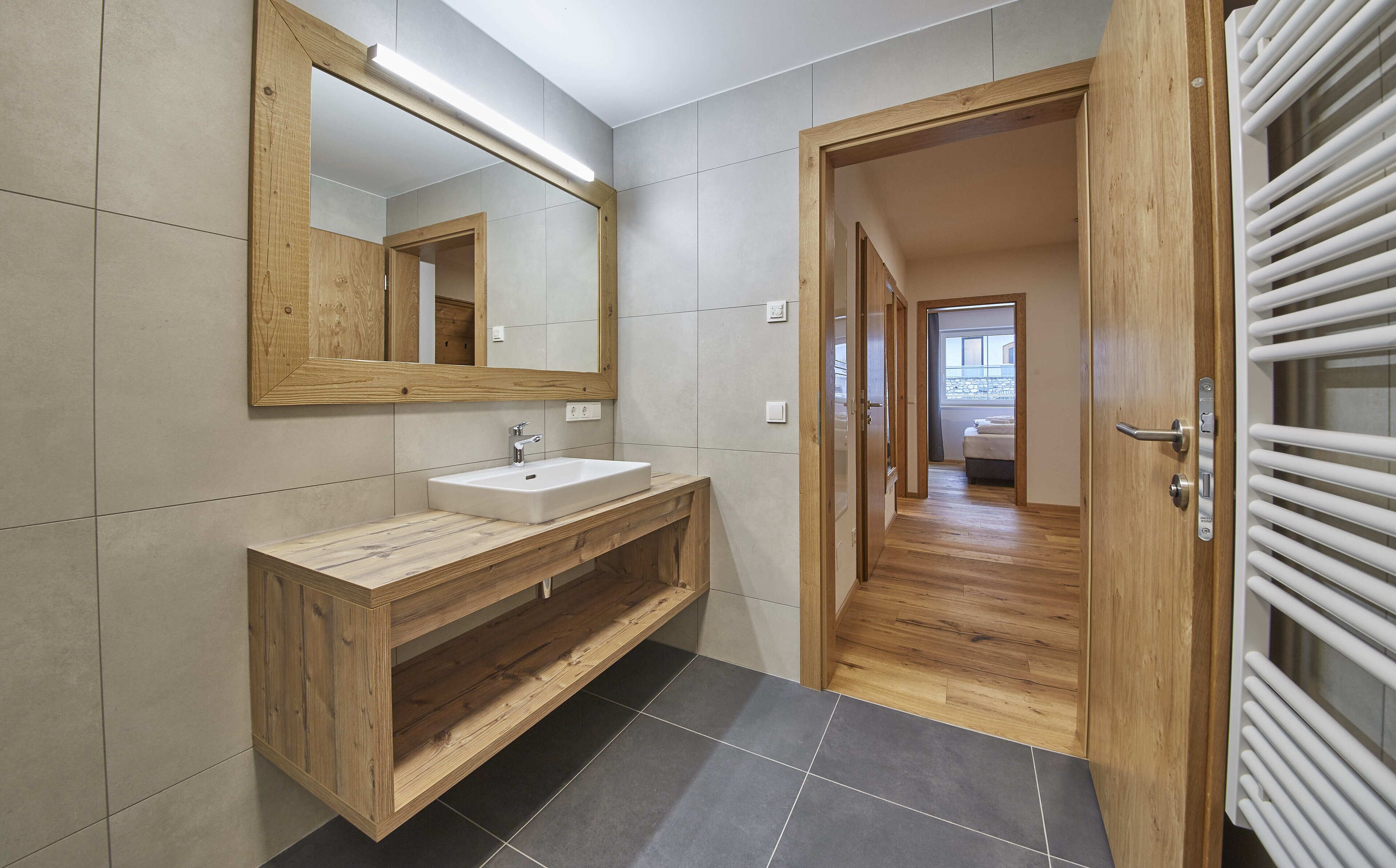 Une salle de bain moderne avec un meuble-lavabo en bois, un grand miroir et une vue sur une chambre à coucher, grâce à une porte ouverte.