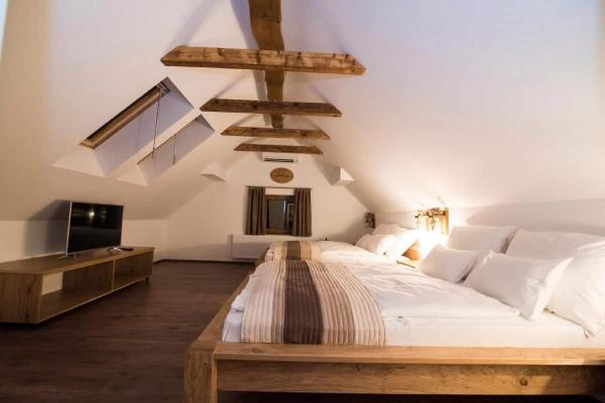 A bedroom with a sloped ceiling, a bed with white bedding, a wooden headboard, a TV on a wooden stand, and a small window.