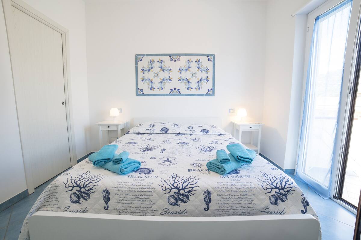 A neatly made bed with a sea-themed bedspread, two bedside tables with lamps, and a window with a view.