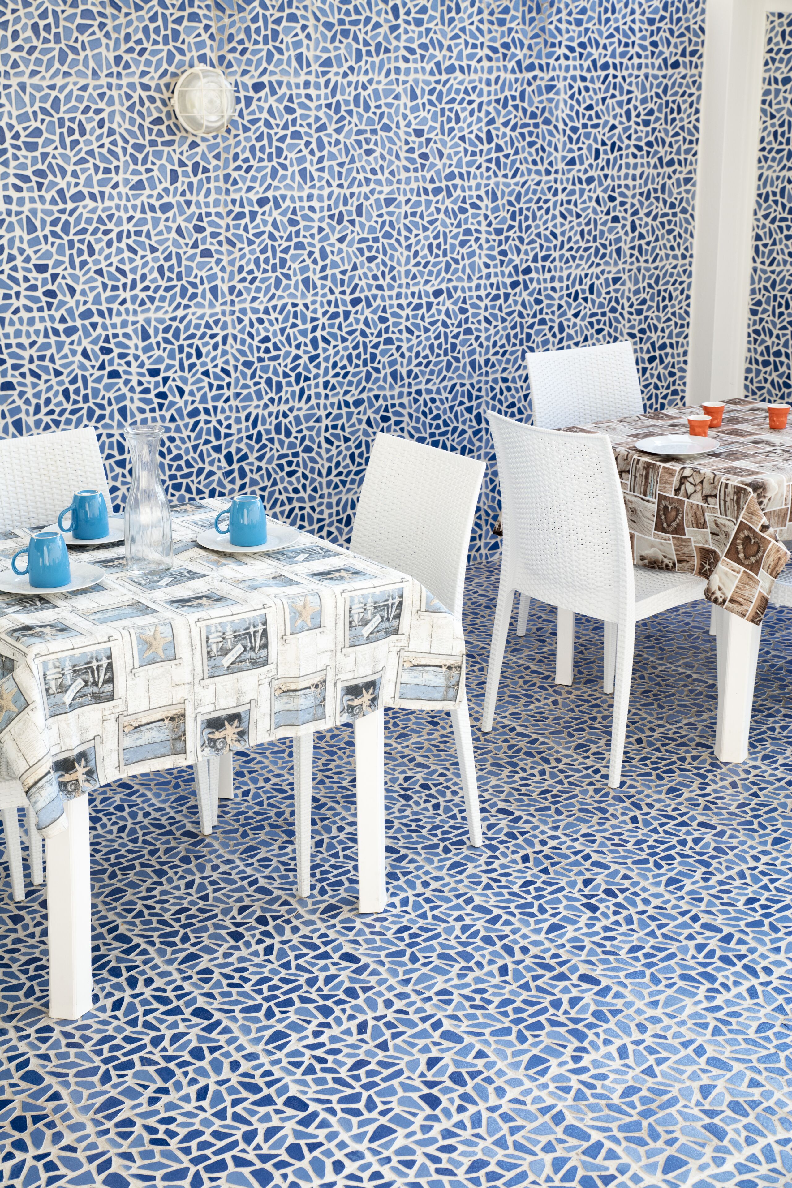 A dining area with mosaic tile flooring and walls, white chairs, and tables with patterned tablecloths.