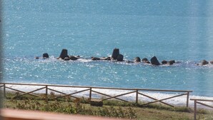 Blick auf die Küste mit einer Reihe von Felsen im Wasser, einem Holzzaun und einem Gebäude mit Balkon im Vordergrund.