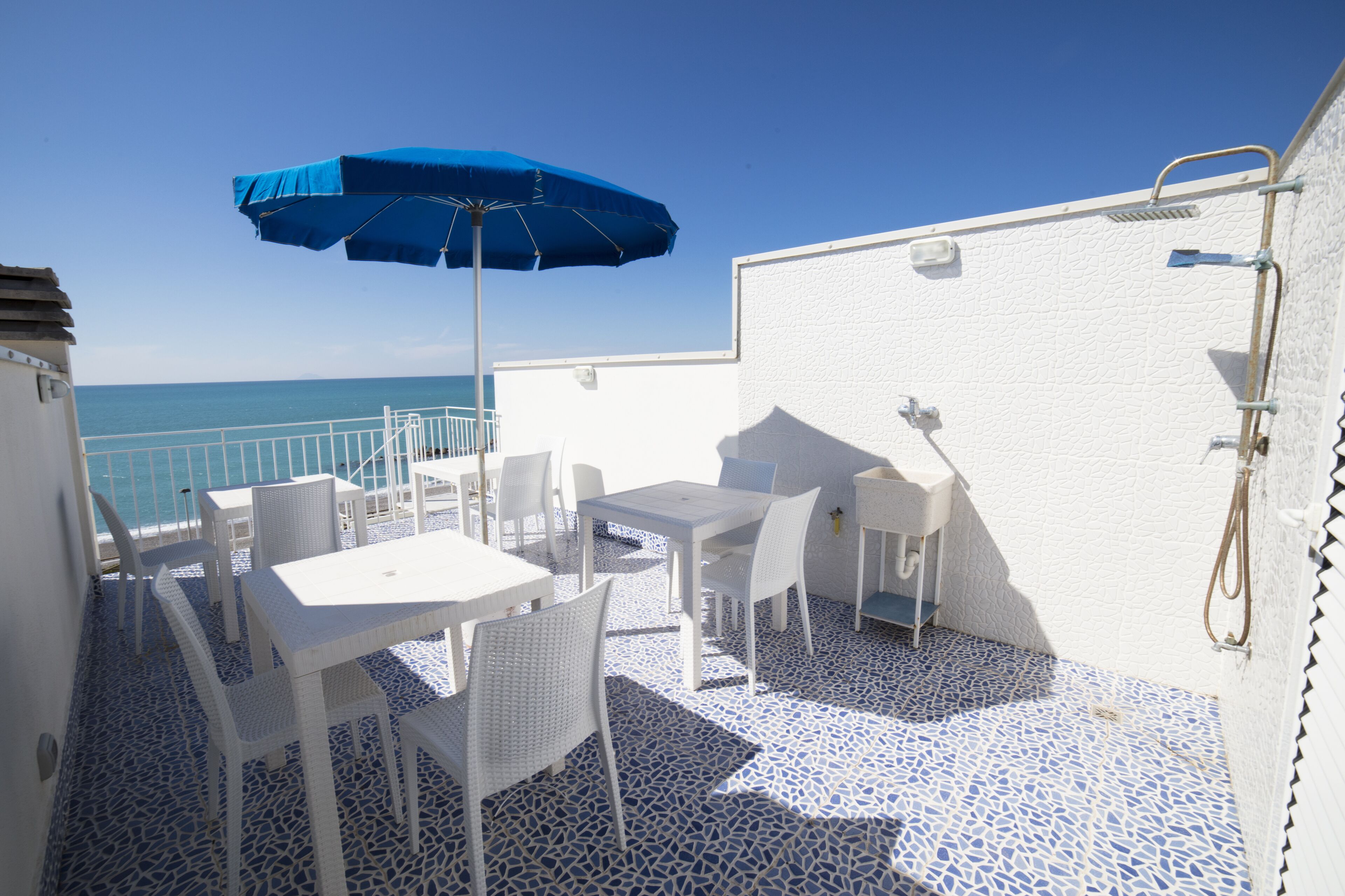 A terrace with white furniture, a blue umbrella, and a view of the sea.