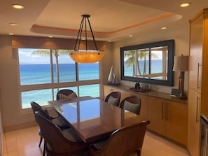 A dining area with a large table and chairs, a hanging light fixture, and a view of the ocean through large windows.