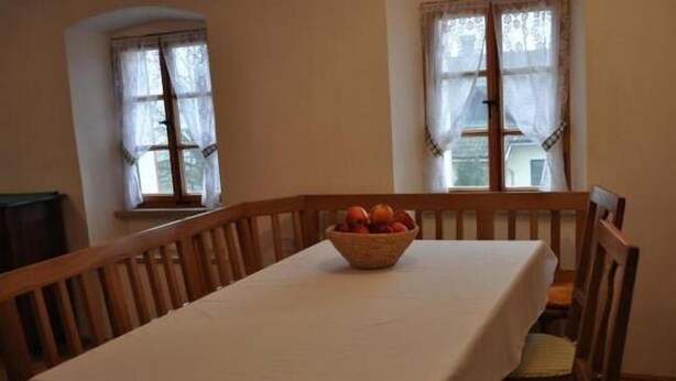 Uma sala de jantar com uma mesa posta para quatro pessoas, duas janelas com cortinas e uma cesta de frutas sobre a mesa.