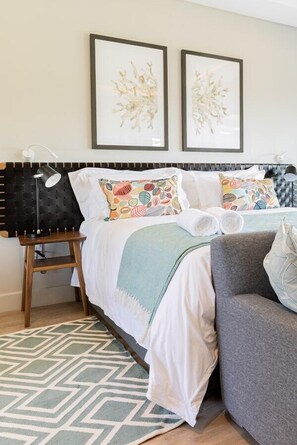 A hotel room with a bed, a bedside table, a chair, and two framed artworks on the wall.