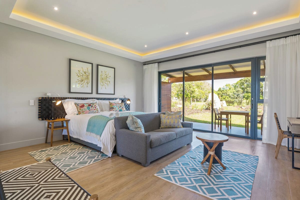 Een moderne slaapkamer met een bed, een grijze bank, een kleine tafel en een balkon met uitzicht op groen.
