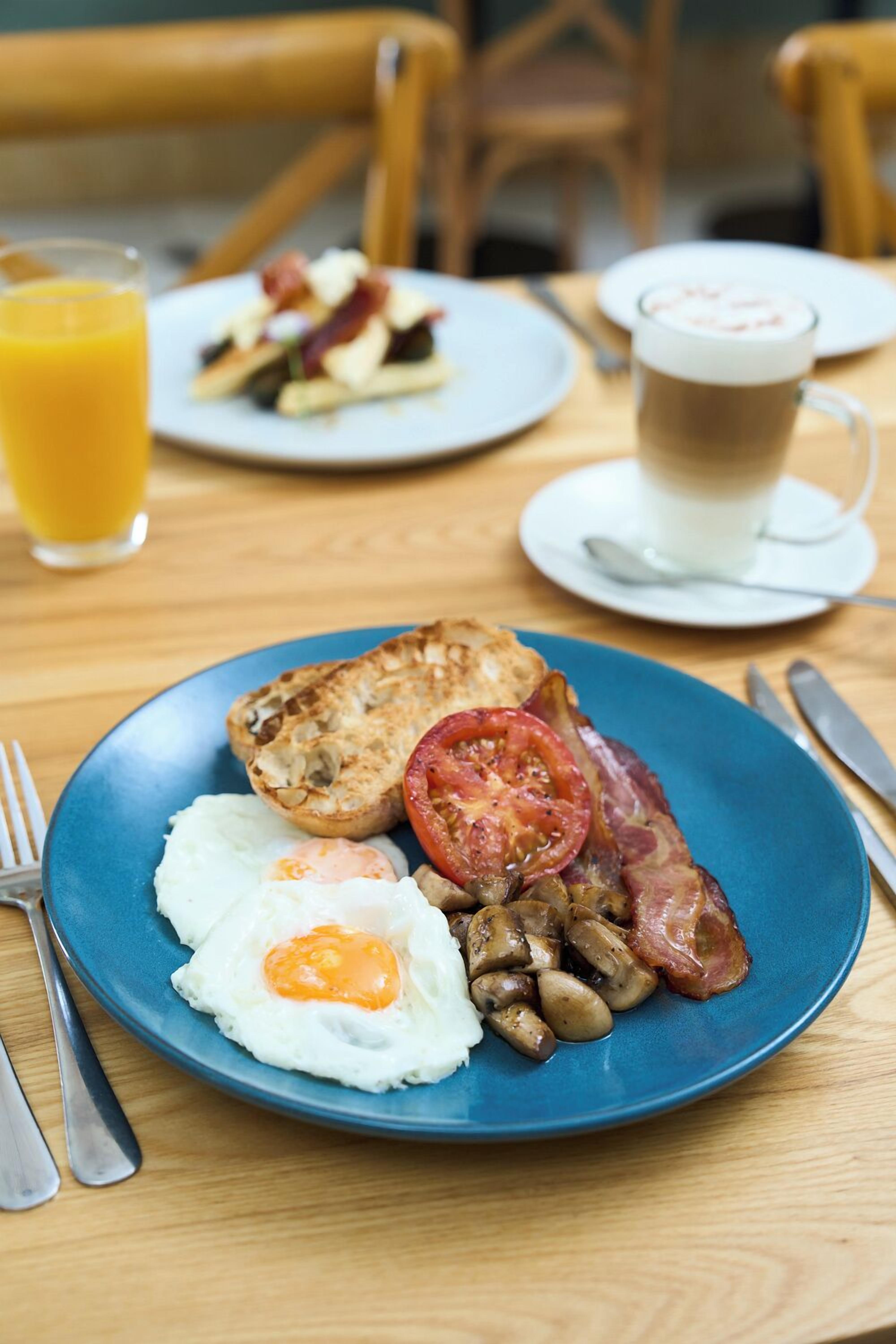Une table dressée pour le petit-déjeuner avec une assiette contenant des œufs, du bacon, des champignons et une tomate, le tout sur une assiette bleue. On y voit également un verre de jus d’orange et une tasse de café posés sur une table en bois.